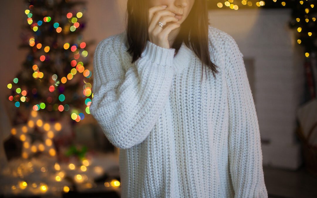 A woman in a white sweater contemplates the holiday depression in front of a Christmas tree and fireplace.